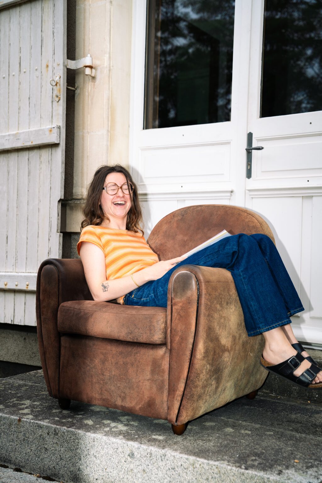 Salomé lit un magazine assise dans un fauteuil de jardin et regarde l’objectif en riant.