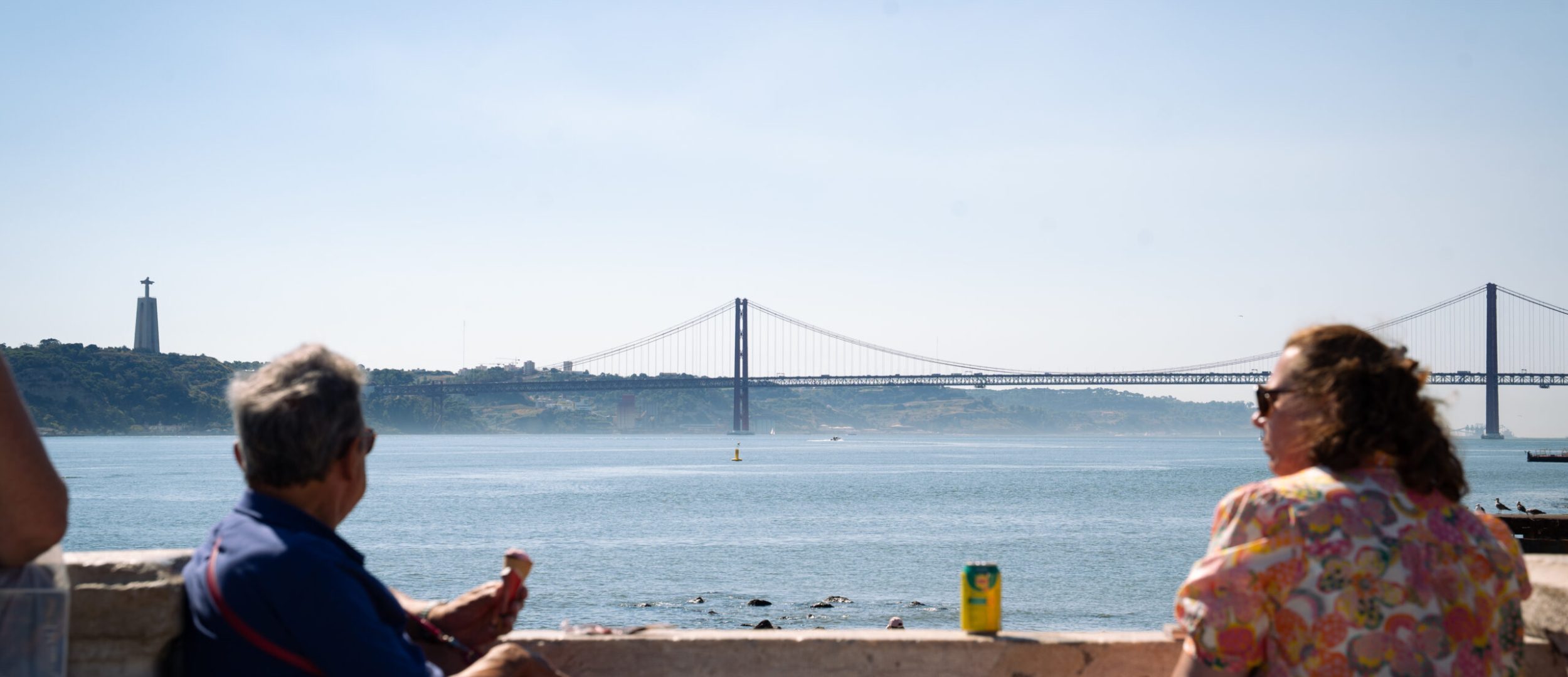 Deux personnes assises sur un banc dégustent une glace tout en admirant le Pont du 25 Avril à Lisbonne.